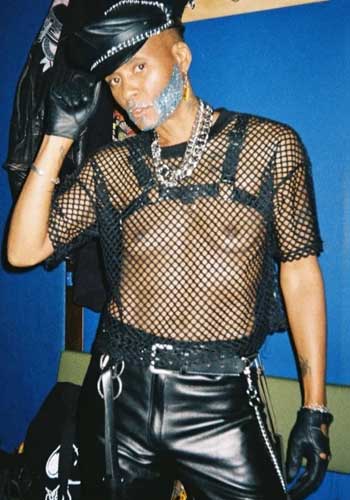 Image: Afro Disiac with silver glitter beard, in leather and mesh shirt outfit, hand tapping on the cap.