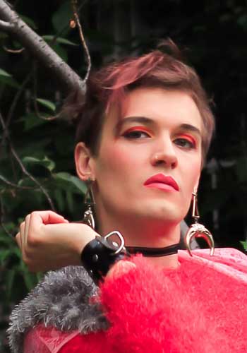 Bild: Brigitte Oytoy mit von unten schauendem Blick in pink rotem Make-Up und Pelzigem Outfit, mit großen silbernen Ohrringen.
